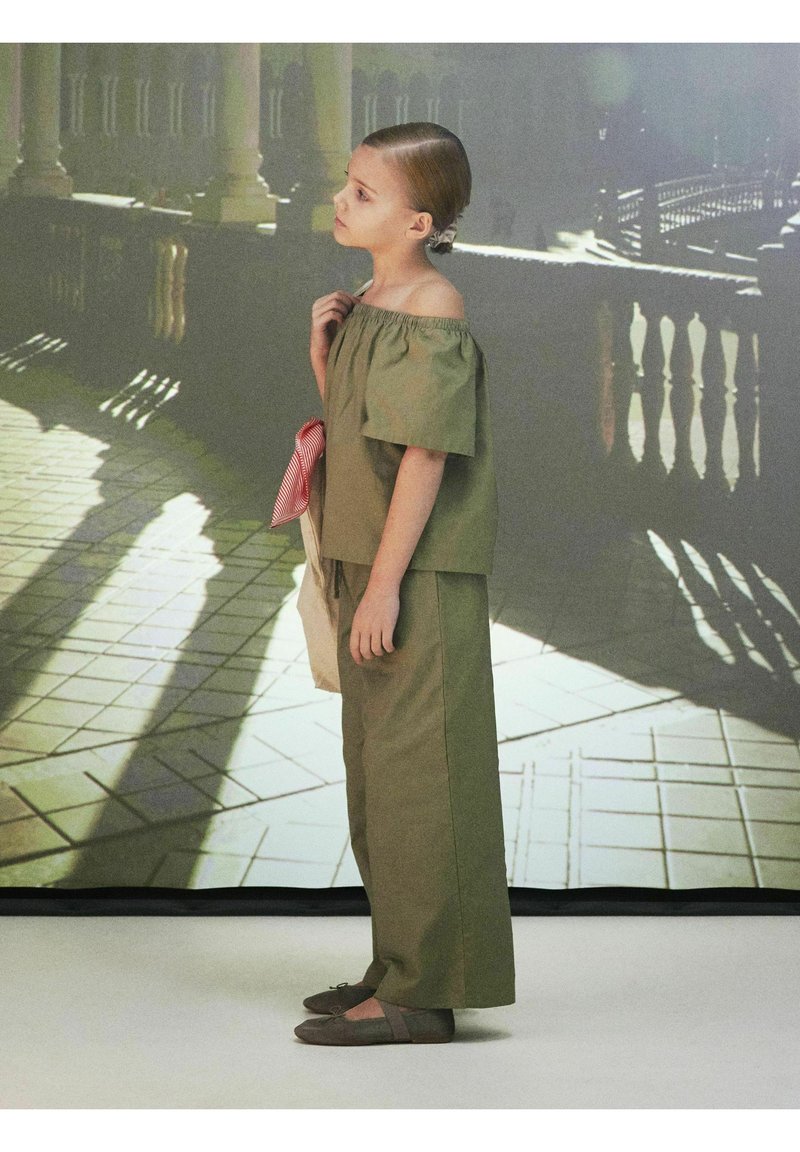 Top off-shoulder verde oliva e pantaloni larghi coordinati; dettaglio a scacchi rosso e bianco; scarpe basse grigie; morbido tessuto di cotone.
