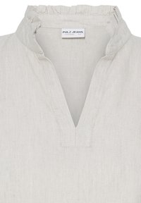 Haut en denim blanc cassé avec un col montant à volants et un décolleté en V, étiquette Pulz Jeans à l'intérieur du col.