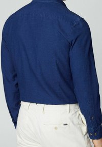 Hombre con camisa de manga larga azul oscuro metida en pantalones de tiro alto color crema con bolsillo trasero abotonado y etiqueta.