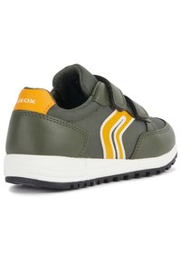 Baskets en cuir vert avec des bandes Velcro, une semelle intermédiaire blanche et des rayures jaunes et blanches distinctives sur le côté. Fini texturé.