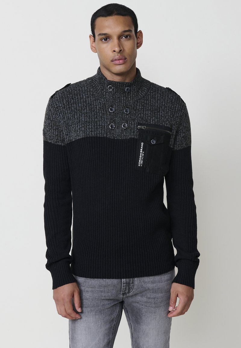 Jersey de punto en negro y gris con textura de canalé, cuello alto, detalle de botones de doble botonadura y un bolsillo en el pecho con cremallera.