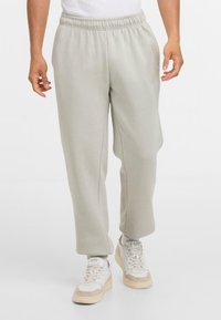 Persoon in lichtgrijze sweatpants en witte sneakers met beige accenten, tegen een eenvoudige witte achtergrond staand.