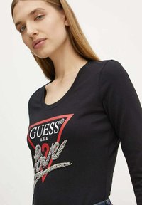 Camiseta negra de manga larga con escote en forma de barco, que presenta un logo triangular rojo y adornos de rhinestone plateados que forman la palabra "Amor".