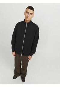 Sort lynlås sweatshirt med høj krave, med sidelommer og ribstrikkede cuffs, parret med løse brune bukser og mørke sneakers.