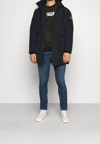 Marineblauwe parka-jas met capuchon, zakken aan de voorkant en trekkoorden. Gedragen over een camouflage-trui, gecombineerd met een blauwe spijkerbroek en witte sneakers.