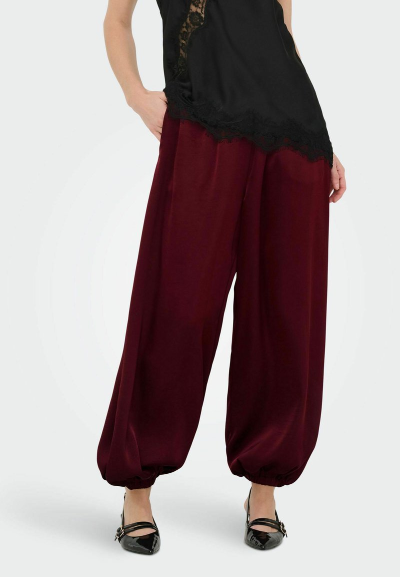 Mannequin portant un pantalon ample en soie bordeaux avec des chevilles froncées, associé à un haut noir orné de dentelle et des escarpins noirs à bouts pointus.