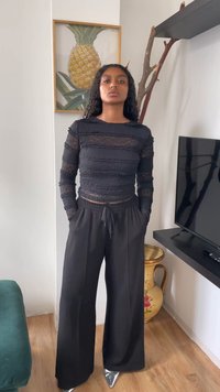 Jovem mulher com cabelo encaracolado, vestindo uma blusa preta transparente com textura, calças largas pretas e sapatos metálicos pontiagudos, em pé numa sala de estar.