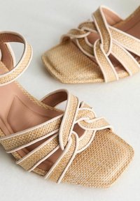 Beige woven sandals with square toe and crossed straps. Soft tan insole with contrasting white trim and texture, highlighting the woven material.