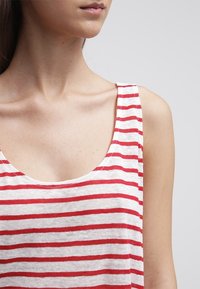 Mladá žena v bílém tílku s vodorovnými červenými pruhy, zobrazená od ramen po těsně pod klíční kostí.