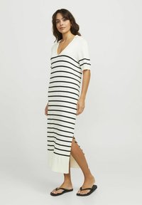 Vestido de punto a rayas en blanco y negro, con escote en V, mangas cortas, aberturas laterales y un corte holgado. Combinado con chanclas negras.