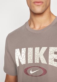 Lys brun bomull t-skjorte med et stort, sprukket hvitt "NIKE"-trykk og en maroon Nike swoosh, med rund hals og korte ermer.