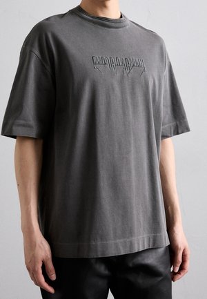 Homme portant un t-shirt ample manches courtes gris foncé avec un motif de texte en relief sur la poitrine, sur fond gris uni.
