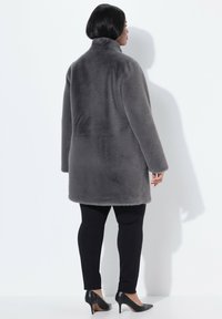 Abrigo de piel sintética gris con cuello alto, mangas largas y corte recto, combinado con pantalones negros ajustados y zapatos de tacón alto negros.