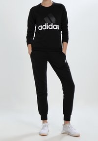 Czarny sweatshirt Adidas z dużym białym logo, w połączeniu z czarnymi zwężanymi dresami. Model ma na sobie białe buty sportowe. Gładka tekstura materiału.