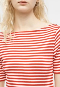 Ξανθός άνθρωπος που φοράει κοντομάνικη μπλούζα με κόκκινα και λευκά ριγέ και ασημένια σκουλαρίκια κρίκους, εμφανιζόμενος από τους ώμους μέχρι τα χείλη.