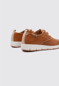 Chaussures décontractées en cuir marron avec des accents beiges, bout rond, semelle blanche et perforations décoratives. Présente une étiquette de marque sur le talon.