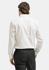 Vit långärmad skjorta med klassisk krage, bakfickor och knappmanschetter, tillsammans med svarta skräddarsydda byxor. Slät textur, skräddarsydd passform.