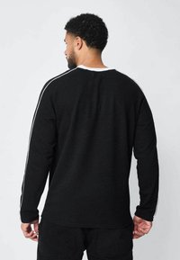 Sweatshirt noir à manches longues avec des rayures blanches sur les côtés, col côtelé et coupe décontractée. La vue arrière montre une texture lisse et des détails de couture subtils.