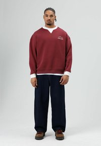 Bordeauxrode trui met een V-hals en witte accenten, gecombineerd met losse donkerblauwe jeans en bruine schoenen, voorzien van subtiele logodetails.