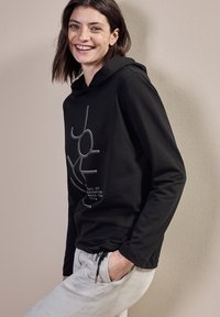 Svart hoodie med dragsko, med en vit grafisk design. Tillsammans med ljusgrå byxor; slätt tyg och avslappnad passform.