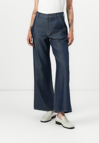 Wide-leg jeans in dark denim, featuring a high waist, front pockets, and a button closure. Paired with cream slip-on shoes.