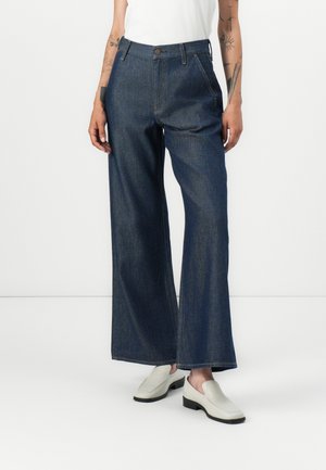 Wide-leg jeans in dark denim, featuring a high waist, front pockets, and a button closure. Paired with cream slip-on shoes.