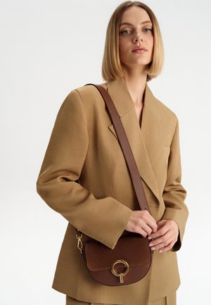 Ruda crossbody rankinė su lygaus odos korpusu, apvaliu auksiniu furnitūros akcentu ir tekstūruota juosta. Nešiojama virš rudos blazerio.