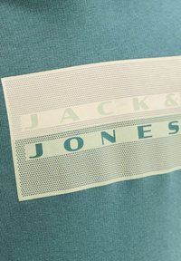 Közeli kép egy zöldesszürke anyagról, amelyen egy téglalap alakú, nyomtatott minta látható, a "JACK & JONES" márkanévvel zöld és sötétzöld betűkkel.