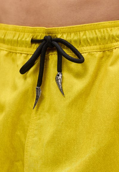 Κίτρινα υφασμάτινα σορτς με μαύρο κορδόνι δεμένο φιόγκο, με μεταλλικά άγκιστρα σε σχήμα μυτερών νυχιών ή δοντιών.