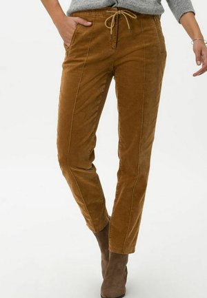 Kobieta ubrana w brązowe spodnie sztruksowe z przednimi kieszeniami i sznurkiem w talii, zestawione z szarym swetrem i brązowymi botkami za kostkę.