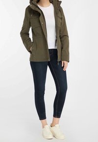 Donna che indossa una giacca verde oliva, una maglietta bianca, jeans skinny scuri e sneakers bianche, in piedi con una mano nella tasca della giacca.