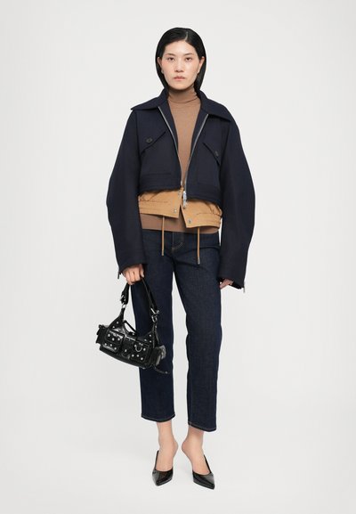 Chaqueta recortada de color azul marino oscuro con gran cuello, cierre frontal y dos botones. Suéter de cuello alto marrón debajo, combinado con jeans oscuros y un bolso negro.