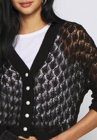 Kvinna iklädd en svart spetstickad kardiagan med pärlknappar över en vit skjorta, som visar midja och arm med avslappnad hållning.