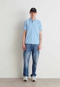 Lyseblå polo skjorte, denimjeans og hvite sneakers. Modellen bærer en svart caps med hvit tekst. Enkelt design og uformell stil.