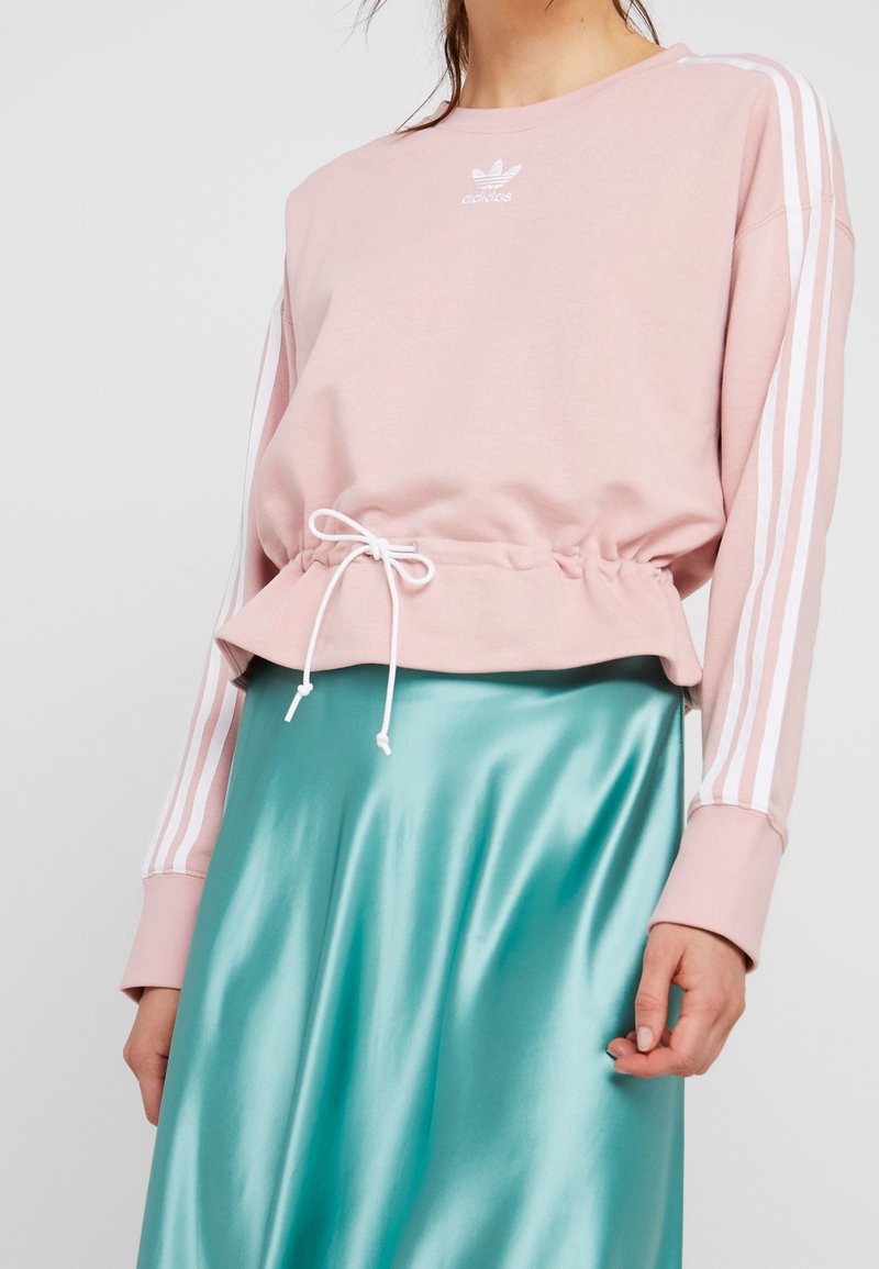 Sweat-shirt rose clair avec une taille froncée et un cordon blanc, présentant des accents blancs à trois rayures sur les manches. Jupe en satin turquoise lisse.
