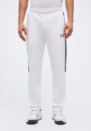 Vyras dėvi baltas EA7 sportines kelnes su juodomis, raudonomis ir žaliomis šoninėmis juostelėmis, derindamas jas su baltos spalvos sportiniais bateliais.