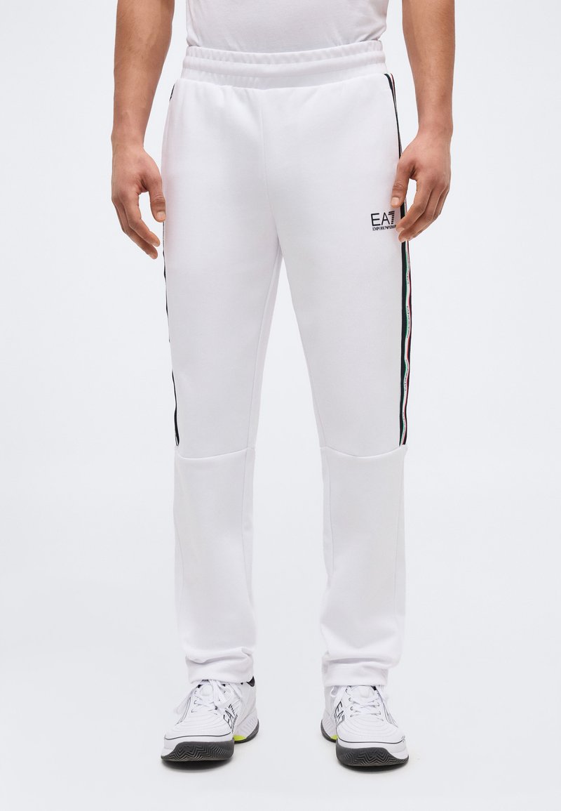 Άνδρας που φοράει λευκή φόρμα EA7 με μαύρες, κόκκινες και πράσινες ρίγες στα πλάγια, συνδυασμένη με λευκά αθλητικά παπούτσια.