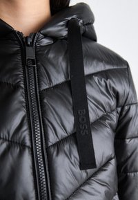 Czarna kurtka puchowa z pikowanym wzorem, wysokim kołnierzem i przednim zamkiem błyskawicznym. Posiada zawieszkę z logo i gładką, refleksyjną teksturę.
