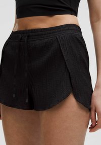 Detailný záber na osobu, ktorá má na sebe čierne štruktúrované športové šortky s viazačkou a čierny cropped top.