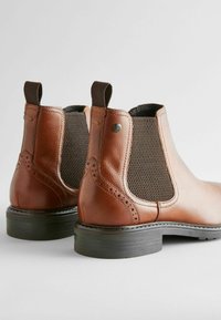 Botas Chelsea de cuero marrón con paneles laterales elásticos, perforaciones decorativas y suelas de goma negra. Textura suave con un diseño elegante.