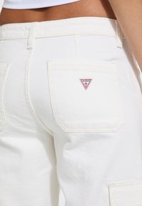 Bijele traperice s visokim strukom. Imaju dva stražnja džepa i mali crveni i bijeli "Guess?" logo na desnom džepu.