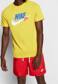 Muž v žlutém tričku Nike s modrým a červeným logem a červených šortkách Nike s bílou šňůrkou a logem na pravé noze.