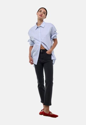 Chemise oversize rayée bleue et blanche, design à boutons, manches retroussées, portée avec un jean noir et des ballerines rouges, pose debout sur fond blanc.