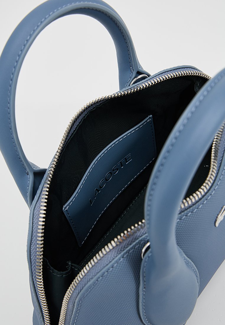 Sac à main synthétique bleu avec une finition texturée, fermeture éclair, deux poignées et une étiquette intérieure arborant "LACOSTE."