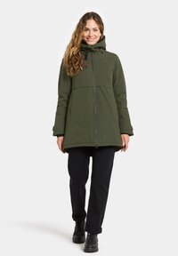 Olijfgroene geïsoleerde parka met rits, verstelbare capuchon en een strakke uitstraling. Gepaard met zwarte broek en veterschoenen.