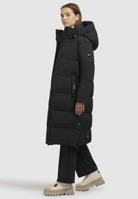 Lang sort pufjakke med hætte, der har lynlås og knaplukning, sidelommer og et quiltet design. Båret med chunky beige fodtøj.