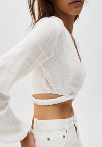 Witte linnen cropped top met lange mouwen, voorzien van een diepe V-hals en een gerimpeld banddetail aan de achterkant. Draagt met witte high-waisted jeans.