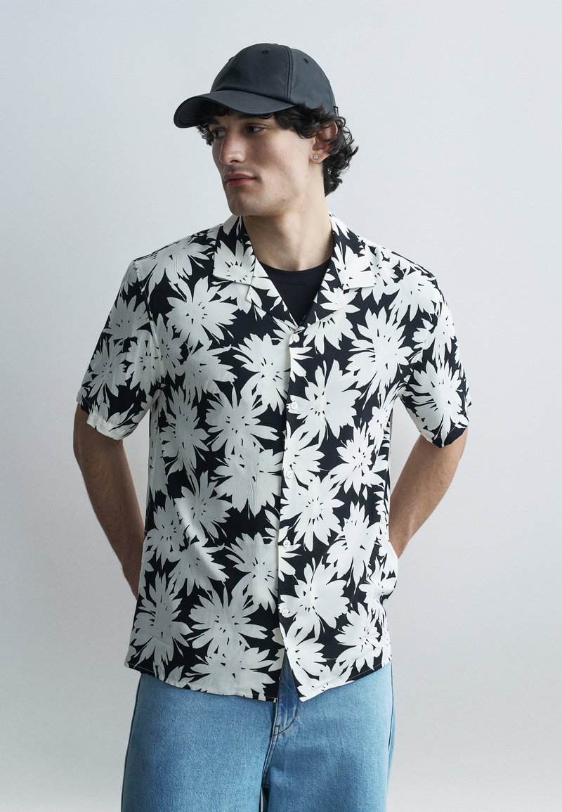 Jeune homme portant une chemise boutonnée à manches courtes à motifs floraux noirs et blancs, un jean bleu clair, une casquette noire et un t-shirt noir, regardant vers la gauche.