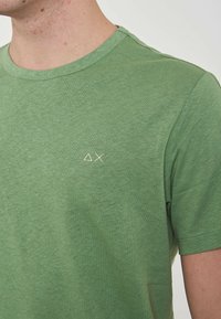 T-shirt verde a maniche corte realizzato in tessuto morbido; presenta un collo rotondo e un piccolo logo chiaro sul petto. Texture liscia.