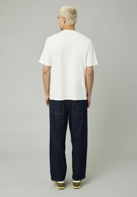 T-shirt en coton blanc à manches courtes, associé à un jean en denim bleu foncé ample. Les chaussures sont grises avec des accents jaunes.